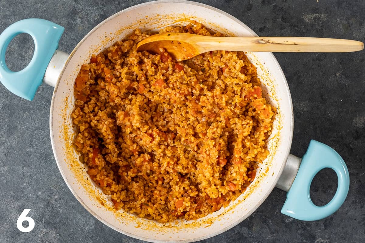 Cooked tomato bulgur pilaf in a pan with a wooden spoon inside it.