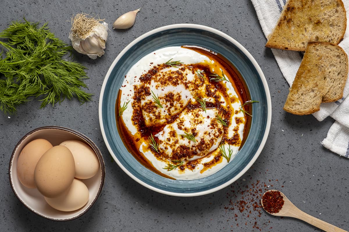 Turkish style poached eggs in a blue bowl, a bunch of fresh dill, eggs with shells in a bowl, garlic cloves, red pepper flakes and toasted bread on the side.
