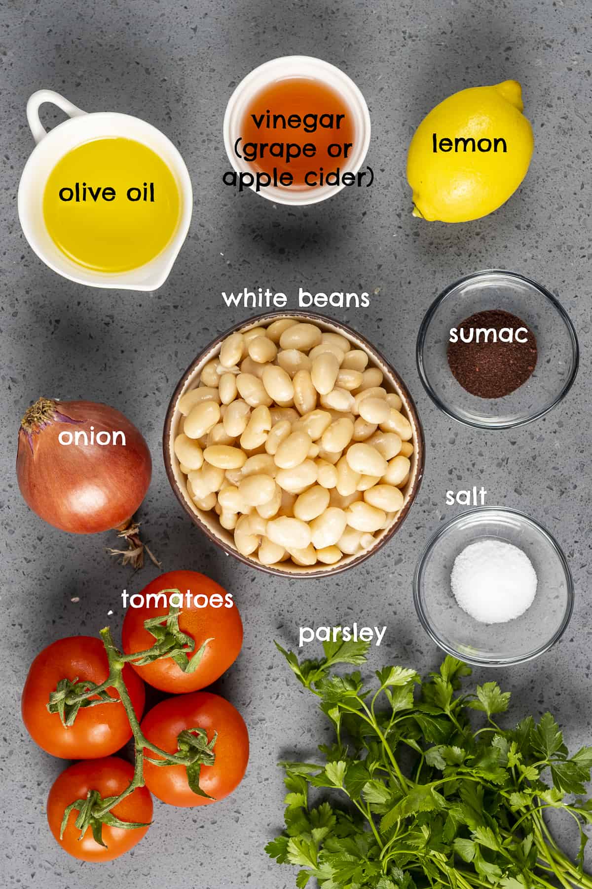 Cooked white beans, tomatoes, olive oil, parsley, lemon, onion, sumac and salt on a grey background.