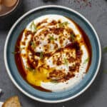 Turkish eggs served in a blue bowl with spicy butter sauce and chopped fresh dill on a dark background.