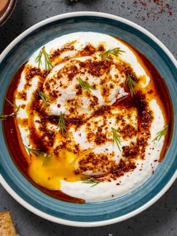 Turkish eggs served in a blue bowl with spicy butter sauce and chopped fresh dill on a dark background.