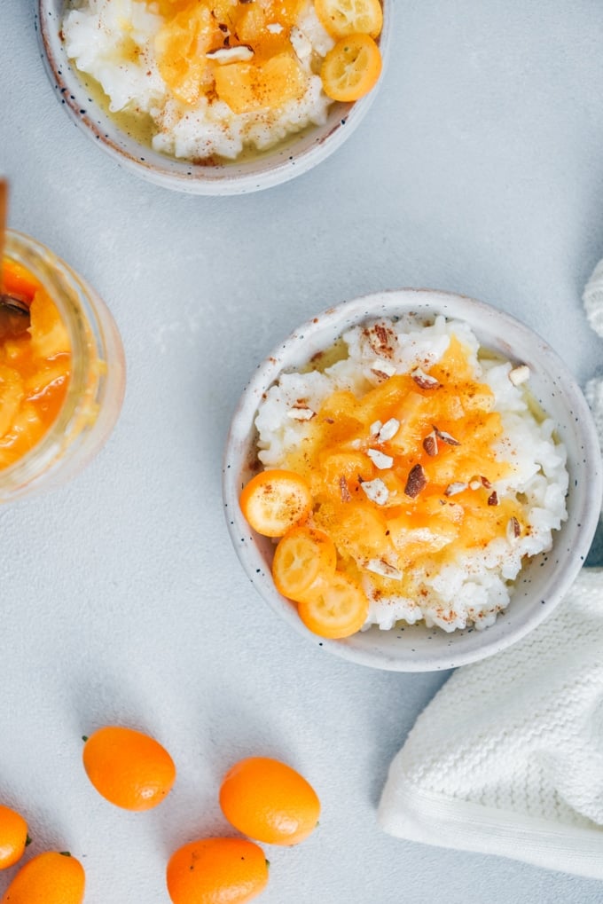 Rice porridge recipe topped with cinnamon, orange jam, kumquats and almonds served in white ceramic bowls.