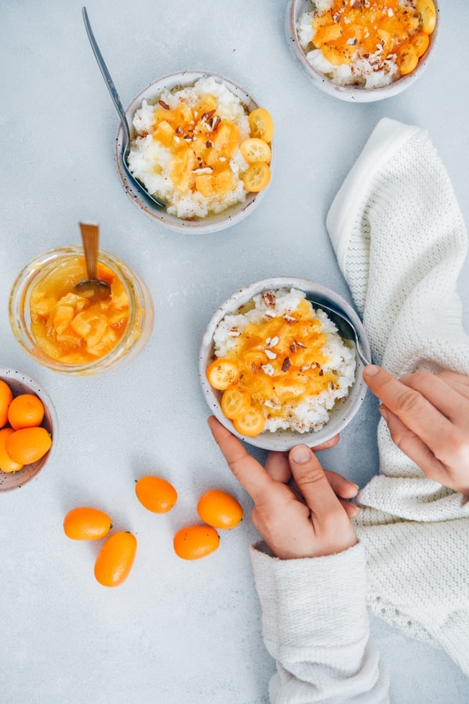 Woman eating rice porridge garnished with orange jam, almonds, cinnamon and kumquats from a white ceramic bowl.