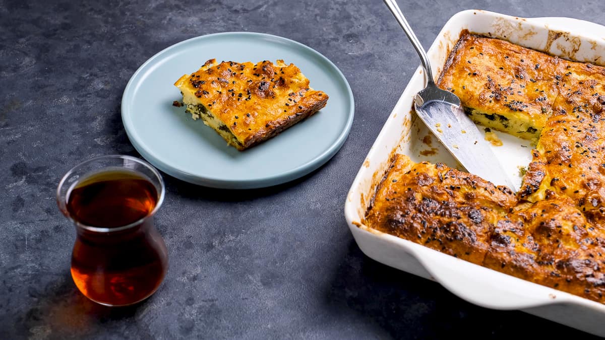 A slice of cheese borek served on a plate, paired with a glass of Turkish tea and the baking pan on the side.