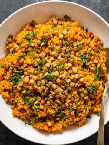 Spicy tomato bulgur pilaf topped with green lentils and parsley in a white bowl with a spoon inside it.