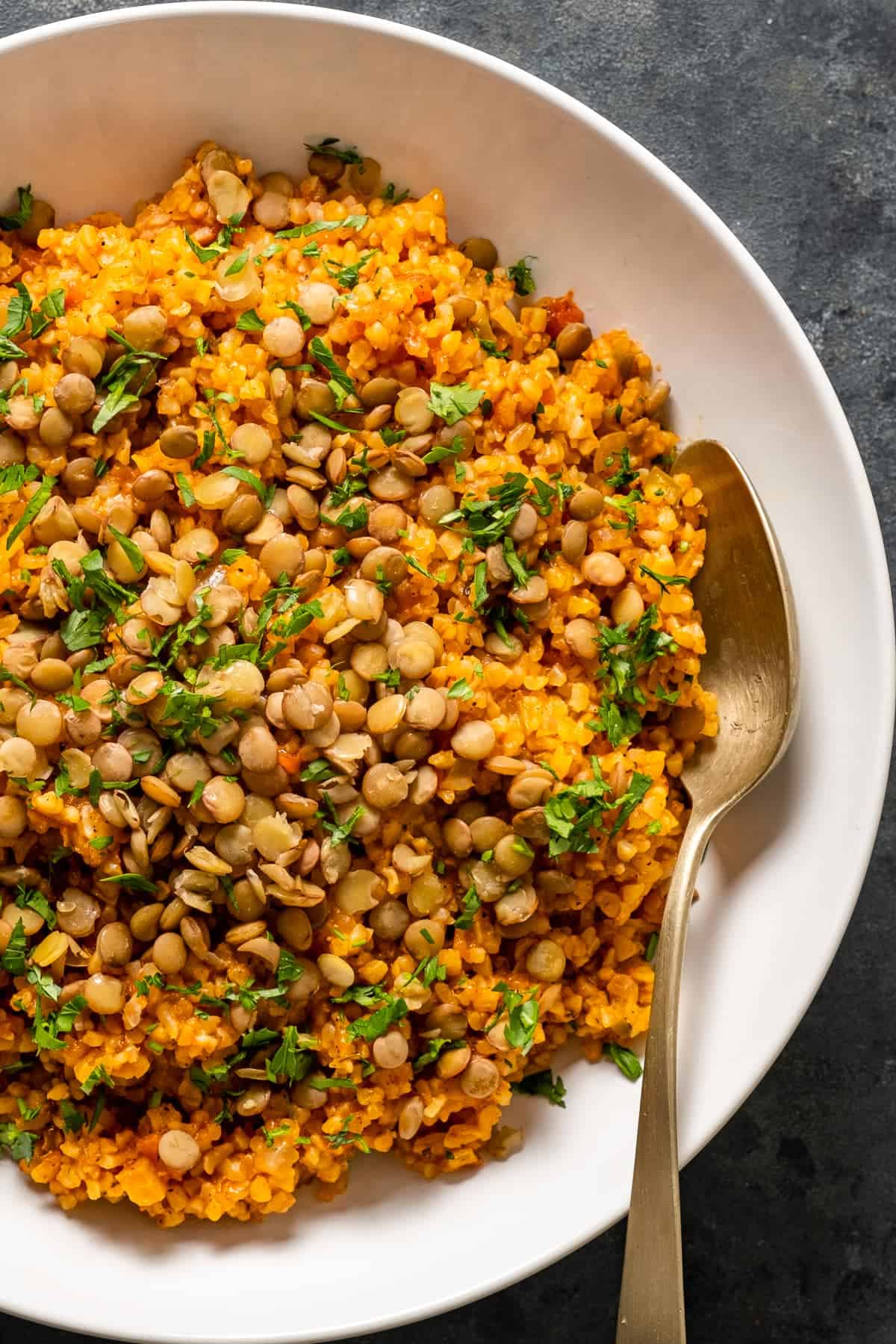 Tomato bulgur pilaf topped with cooked green lentils and chopped parsley in a white bowl with a spoon inside it.