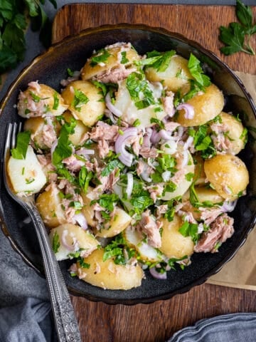Tuna potato salad served in a black bowl and a fork inside, lemon halves, parsley and black pepper on the side.