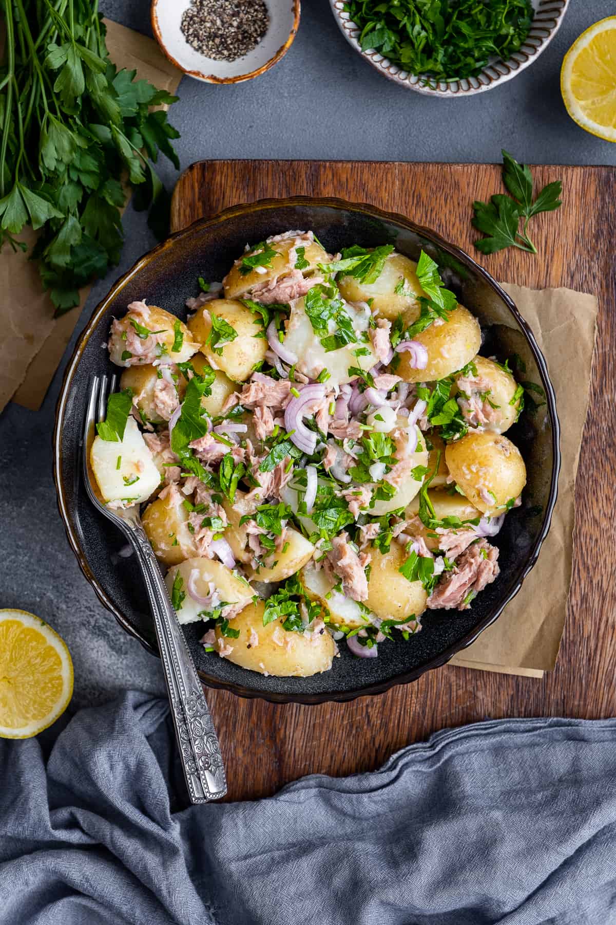 Tuna potato salad served in a black bowl and a fork inside, lemon halves, parsley and black pepper on the side.