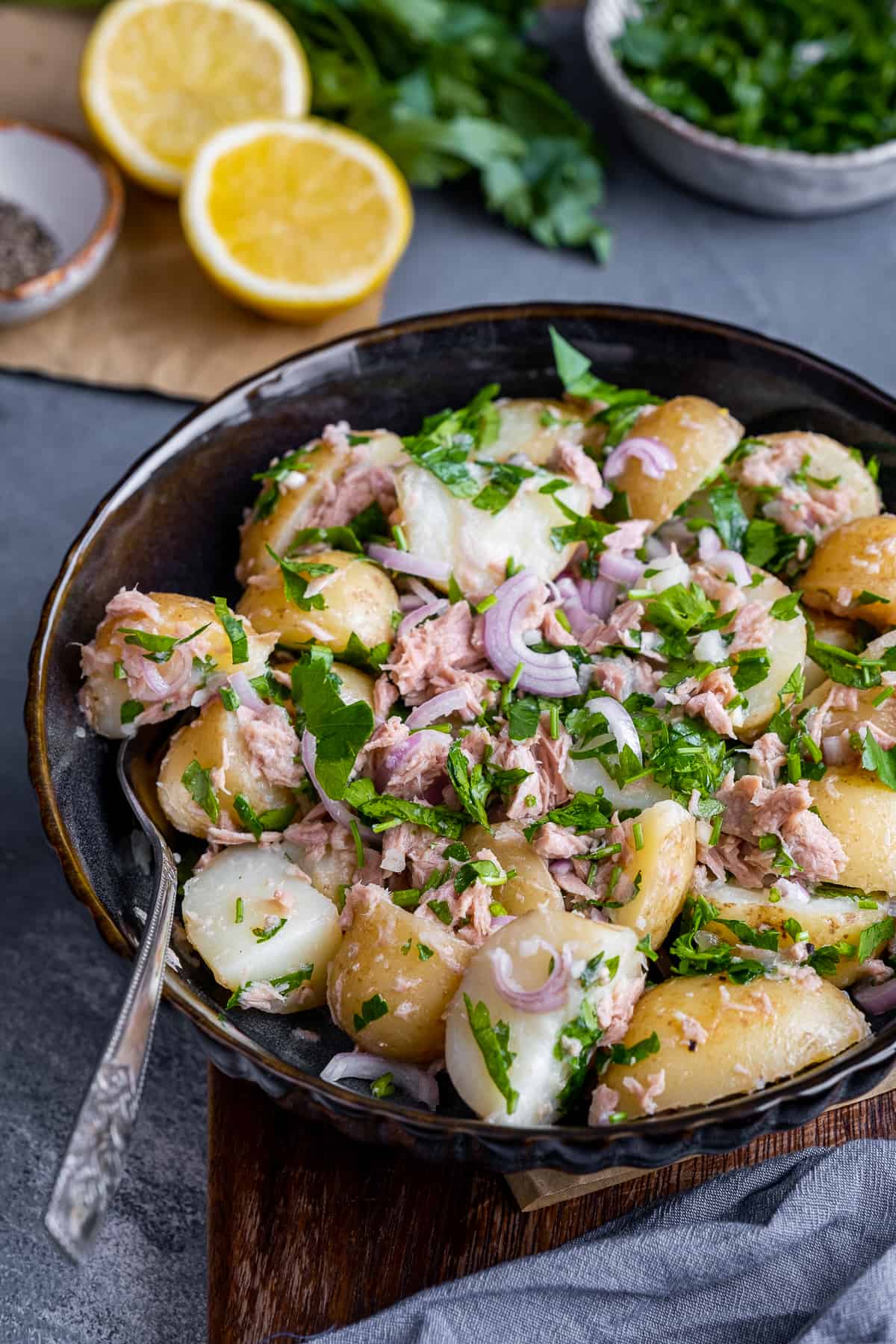 Potato salad with tuna, onions and parsley in a black bowl, a fork inside it, lemon halves and parsley behind it.