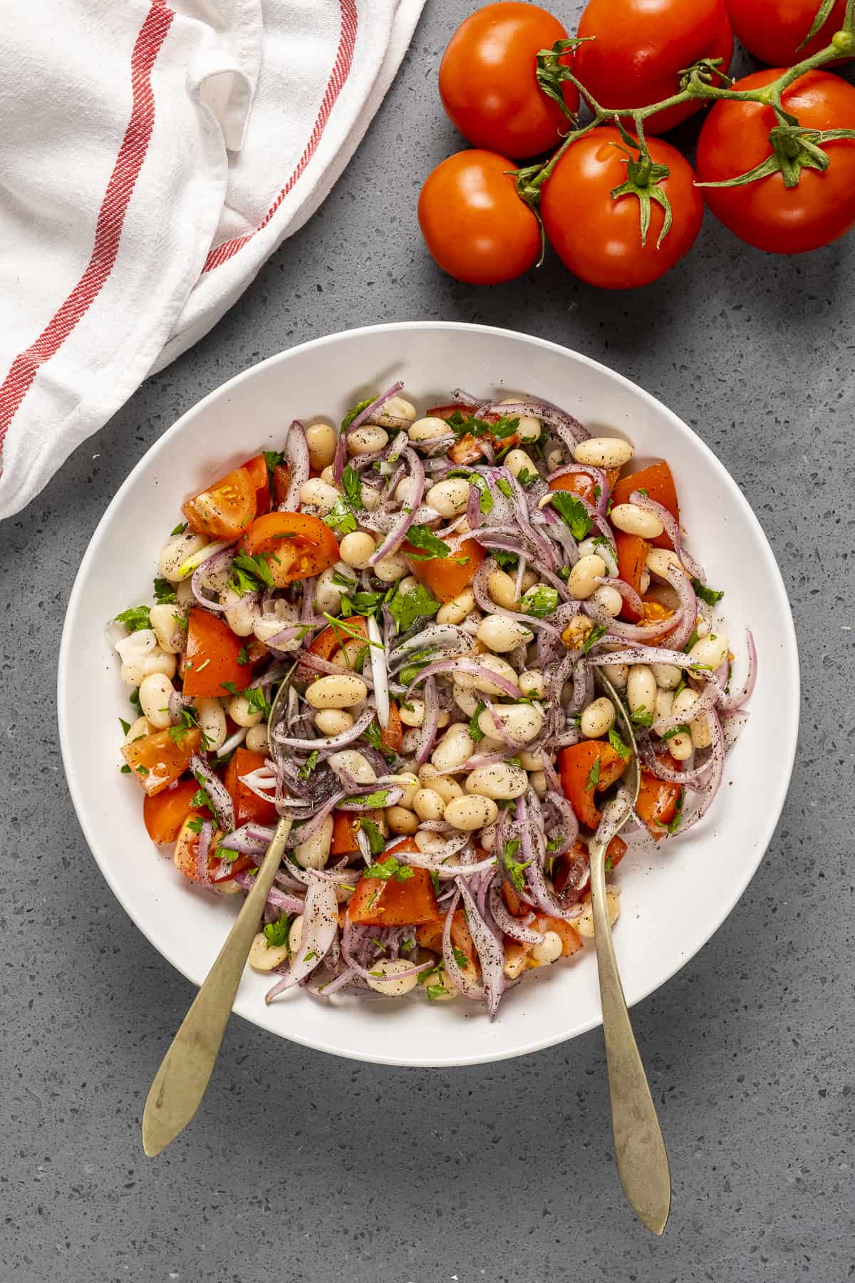 Bean salad with tomatoes and onions in a white bowl and two spoons in it.