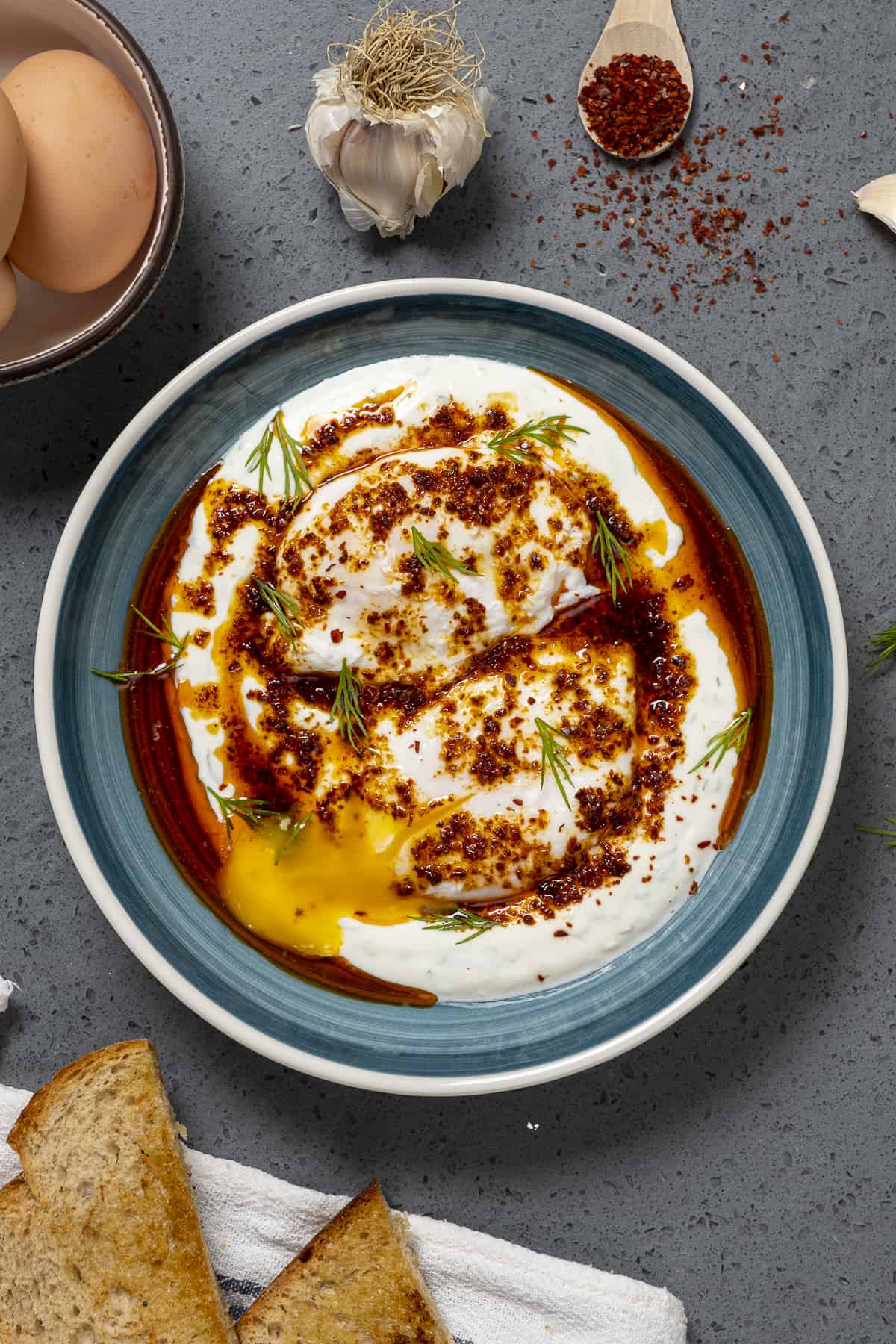 Poached eggs on Greek yogurt served with spiced butter sauce in a blueish bowl. Garlic cloves, toasted bread, pepper flakes and eggs in shell on the side.