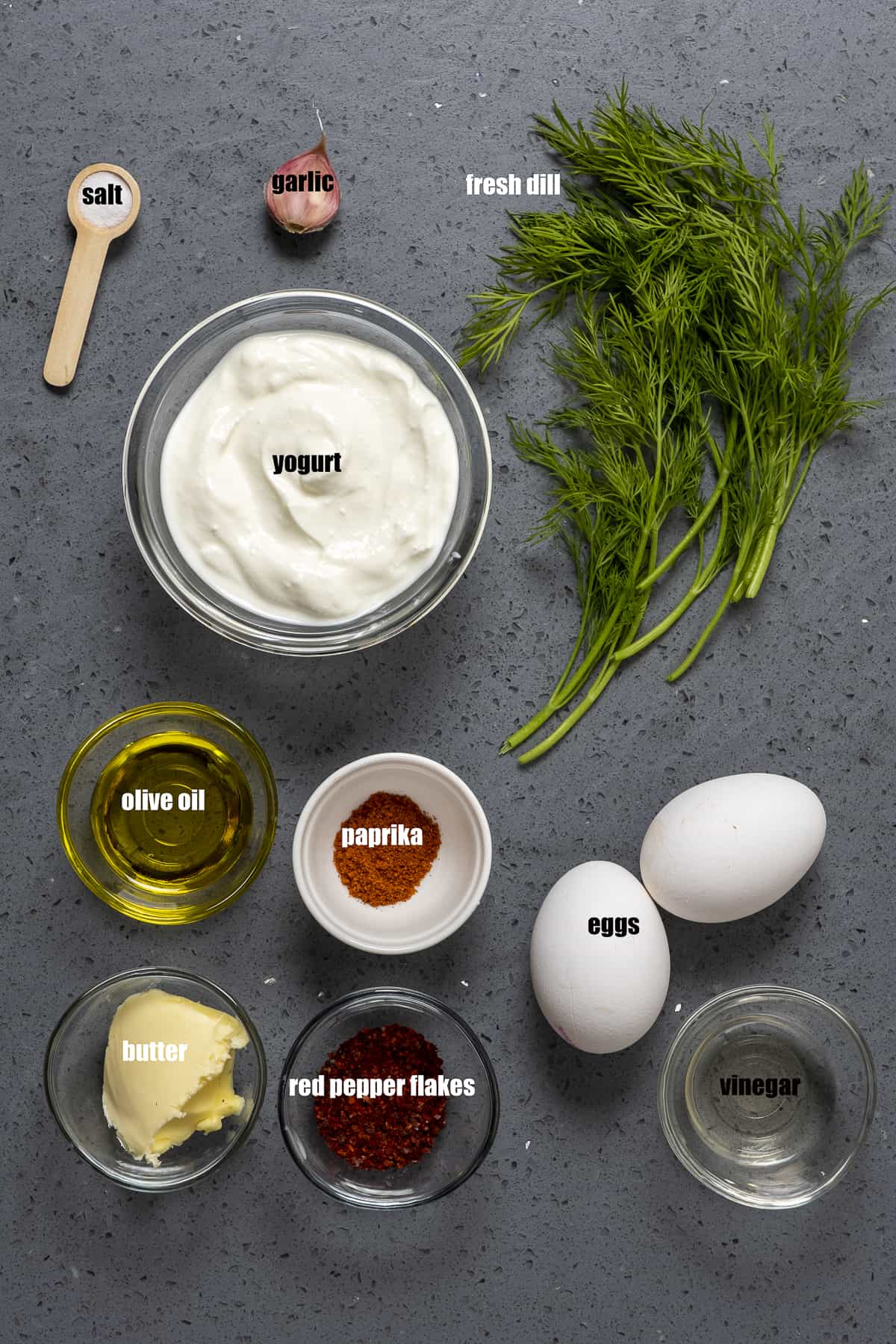 Eggs, Greek yogurt, fresh dill, butter, olive oil, red pepper flakes, paprika, garlic clove, salt in a teaspoon on a dark background.