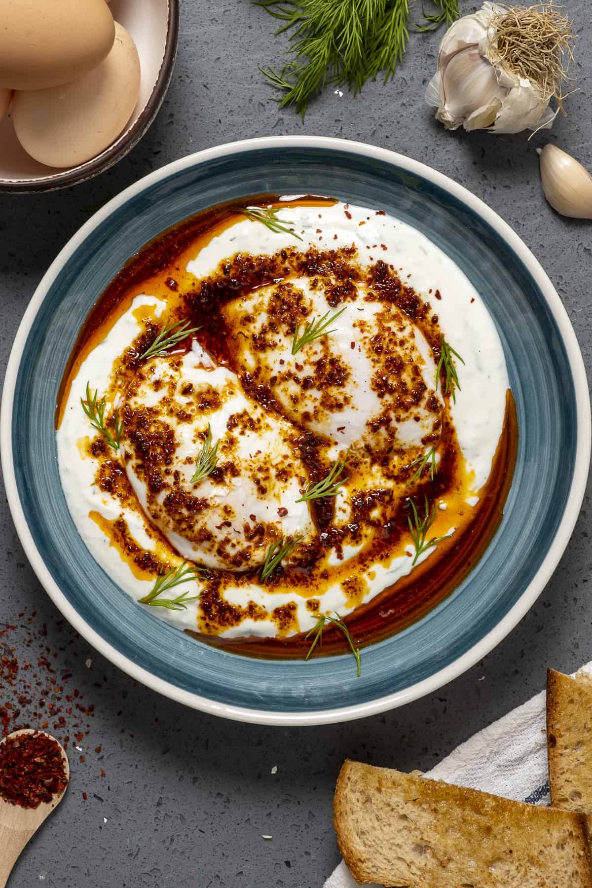 Poached eggs on Greek yogurt served with spiced butter sauce in a blueish bowl. Garlic cloves, toasted bread, fresh dill, pepper flakes and eggs in shell on the side.