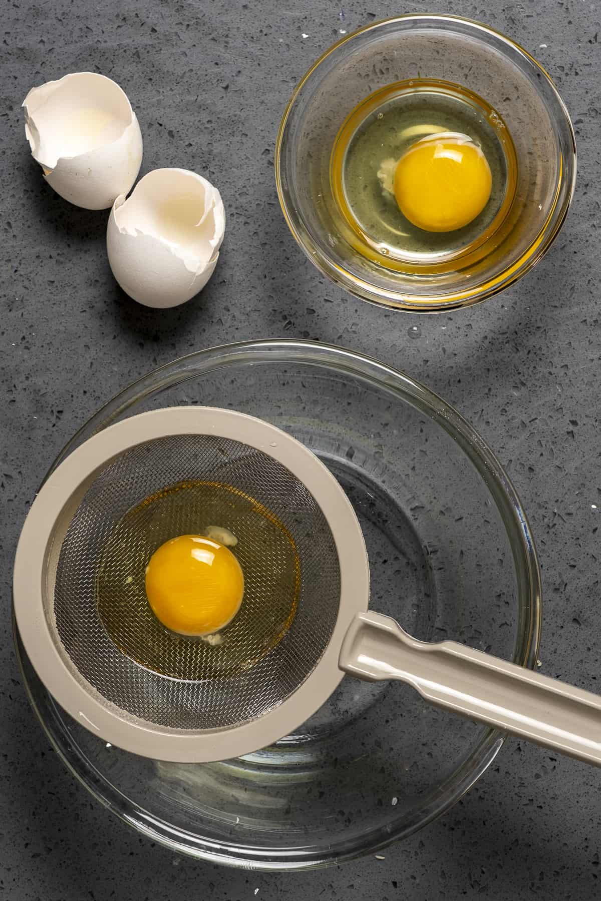 One egg is cracked into a finemesh strainer placed over a glass bowl and another egg in a small bowl and egg shells on the side.