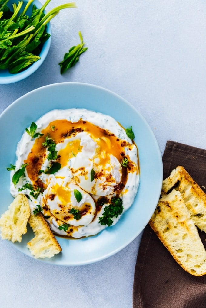 Turkish eggs served in a blue bowl drizzled with a butter sauce accompanied by spinach and toasted bread.