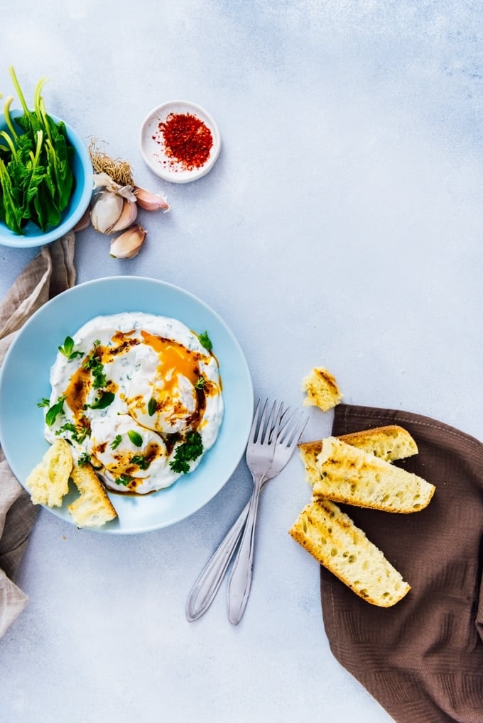 Turkish eggs served in a blue bowl drizzled with a butter sauce accompanied by spinach, red pepper flakes and toasted bread.
