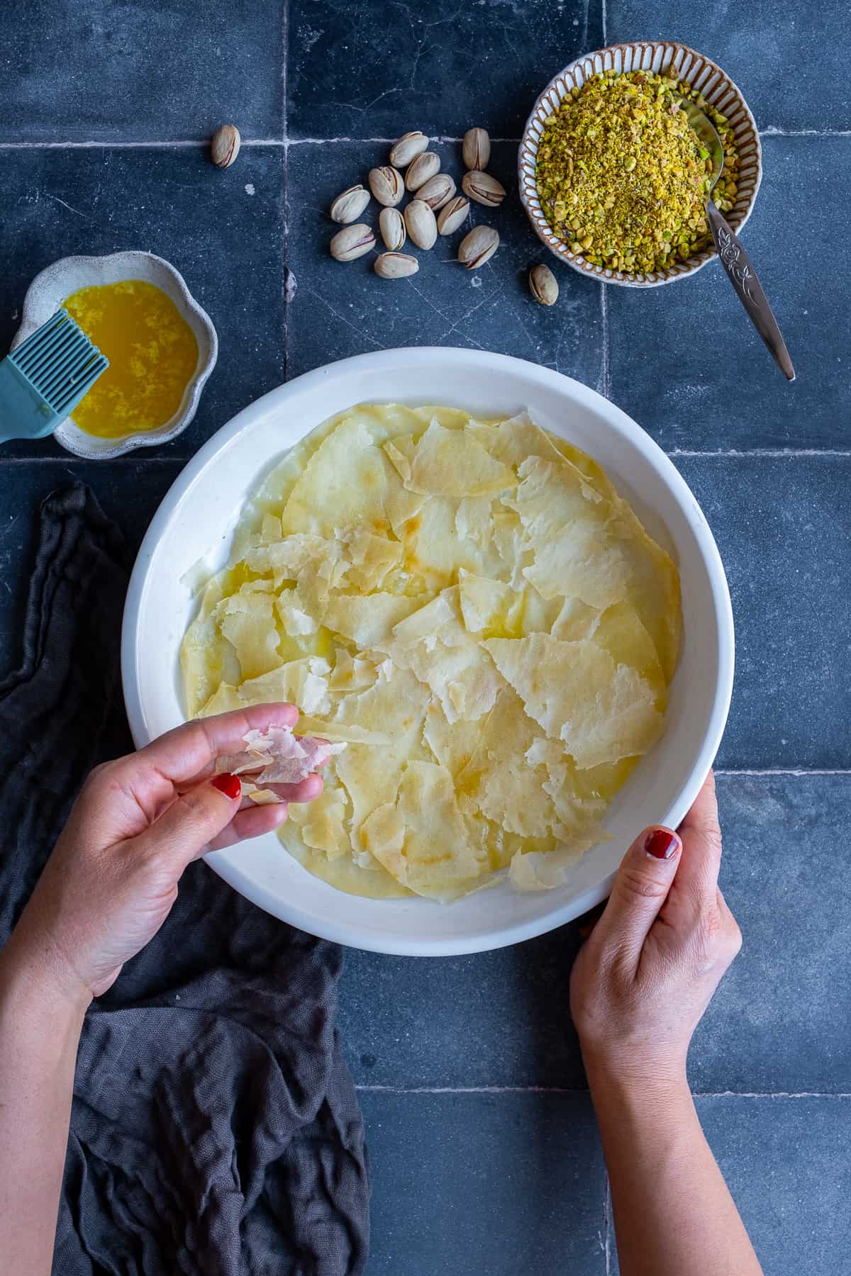 Hands placing torn phyllo sheets in a round baking pan, melted butter and a brush, pistachios and chopped pistachios on the side.