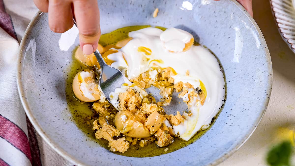 Hand mixing cooked egg yolks and Greek yogurt with a fork in a bowl.