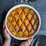 Hands holding a round baking pan full of baklava dessert on a grey background.