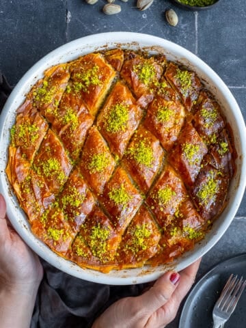 Hands holding a round baking pan full of baklava dessert on a grey background.