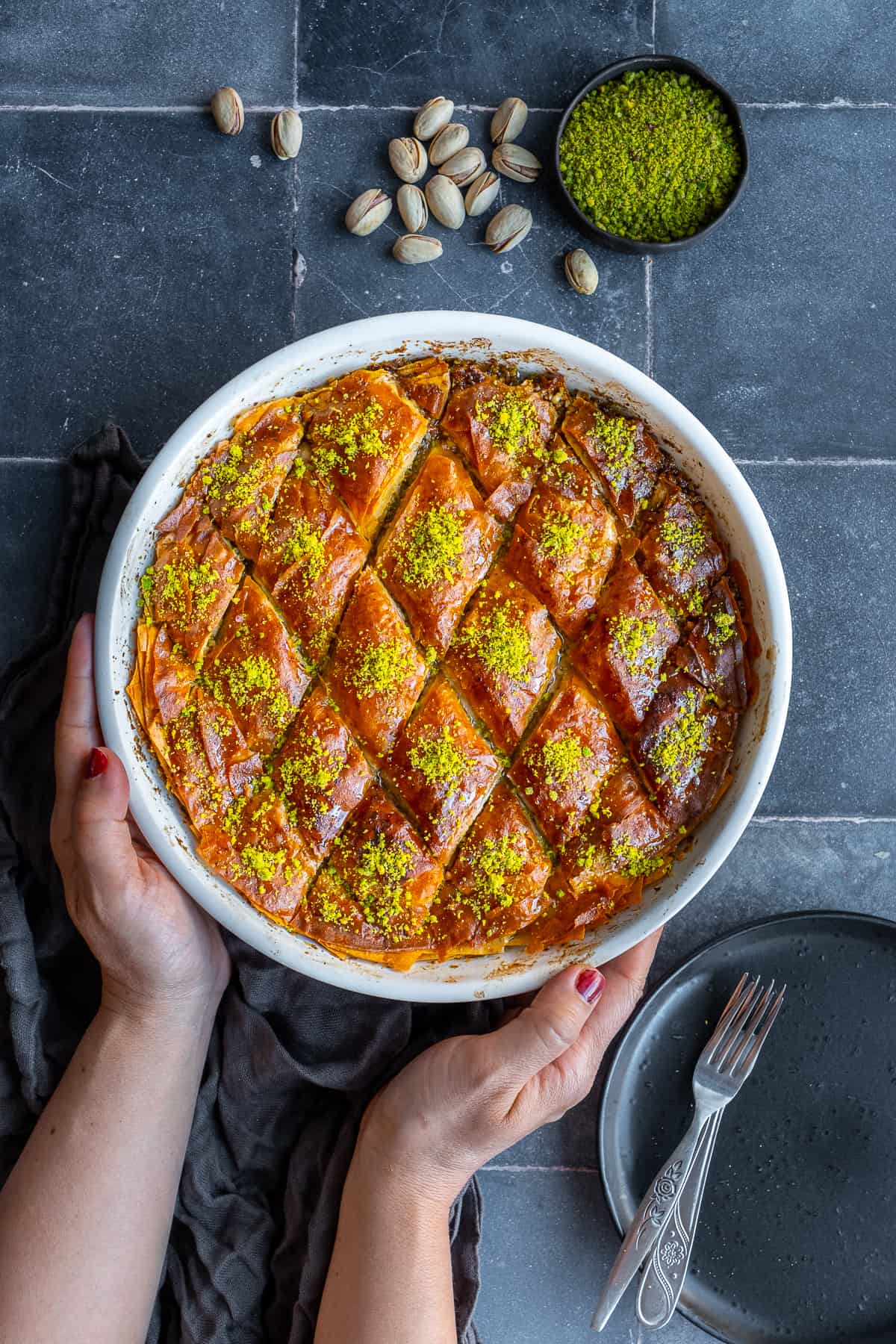 Woman hands holding baklava dessert in a round baking pan, pistachios and ground pistachio on the side.