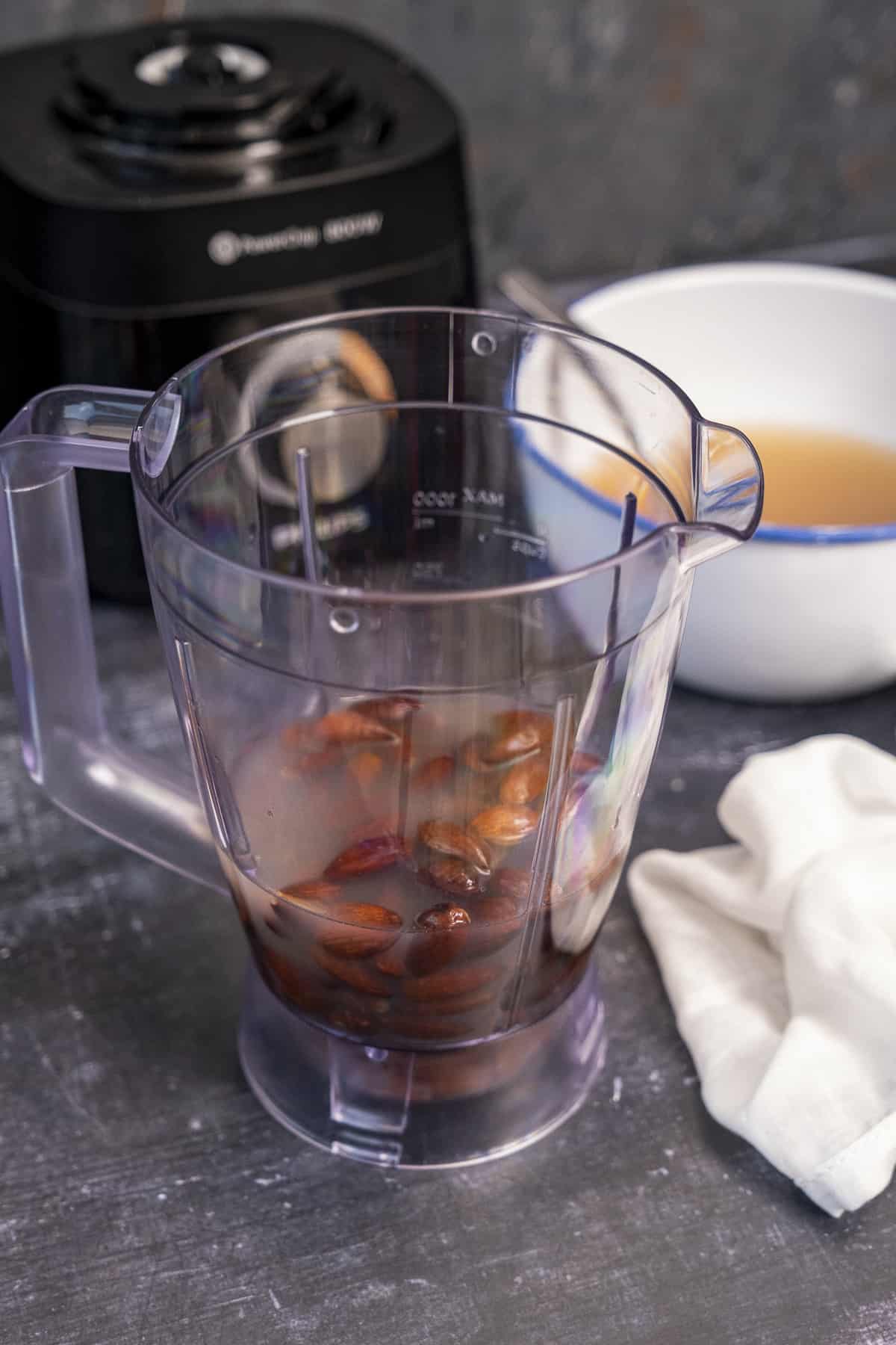 Roasted and soaked almonds in a blender, soaking water in a white bowl and a white muslin are on the side.