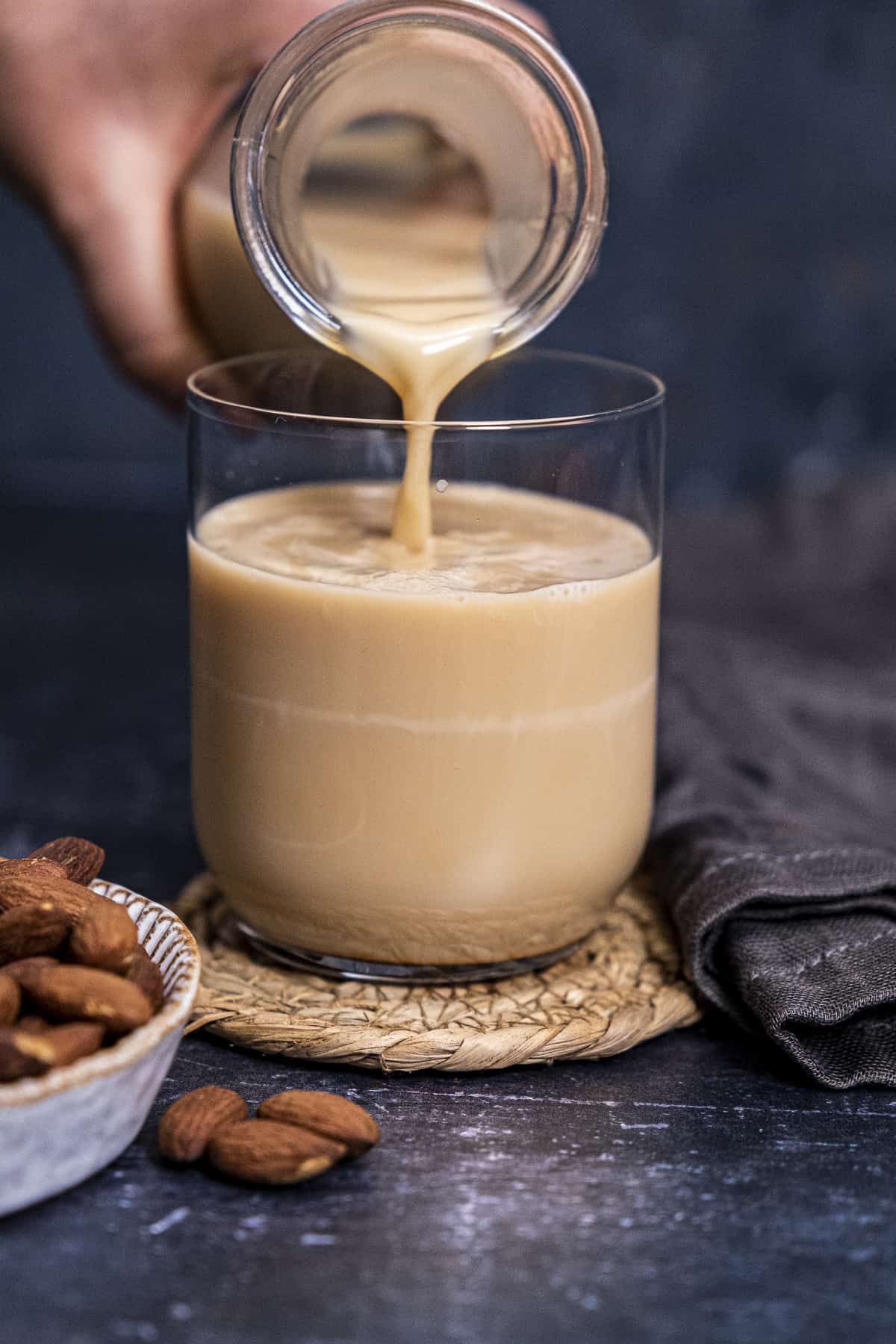 A hand pouring roasted almond milk into a glass and whole almonds on the side.