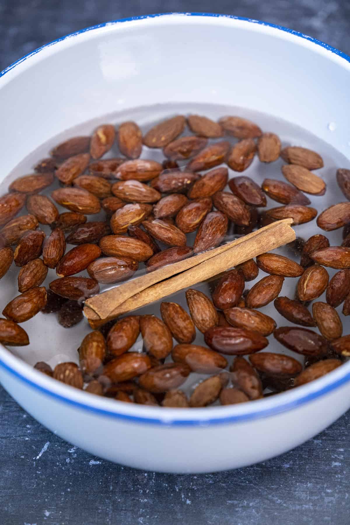 Whole almonds and a cinnamon stick soaked in water in a white bowl.