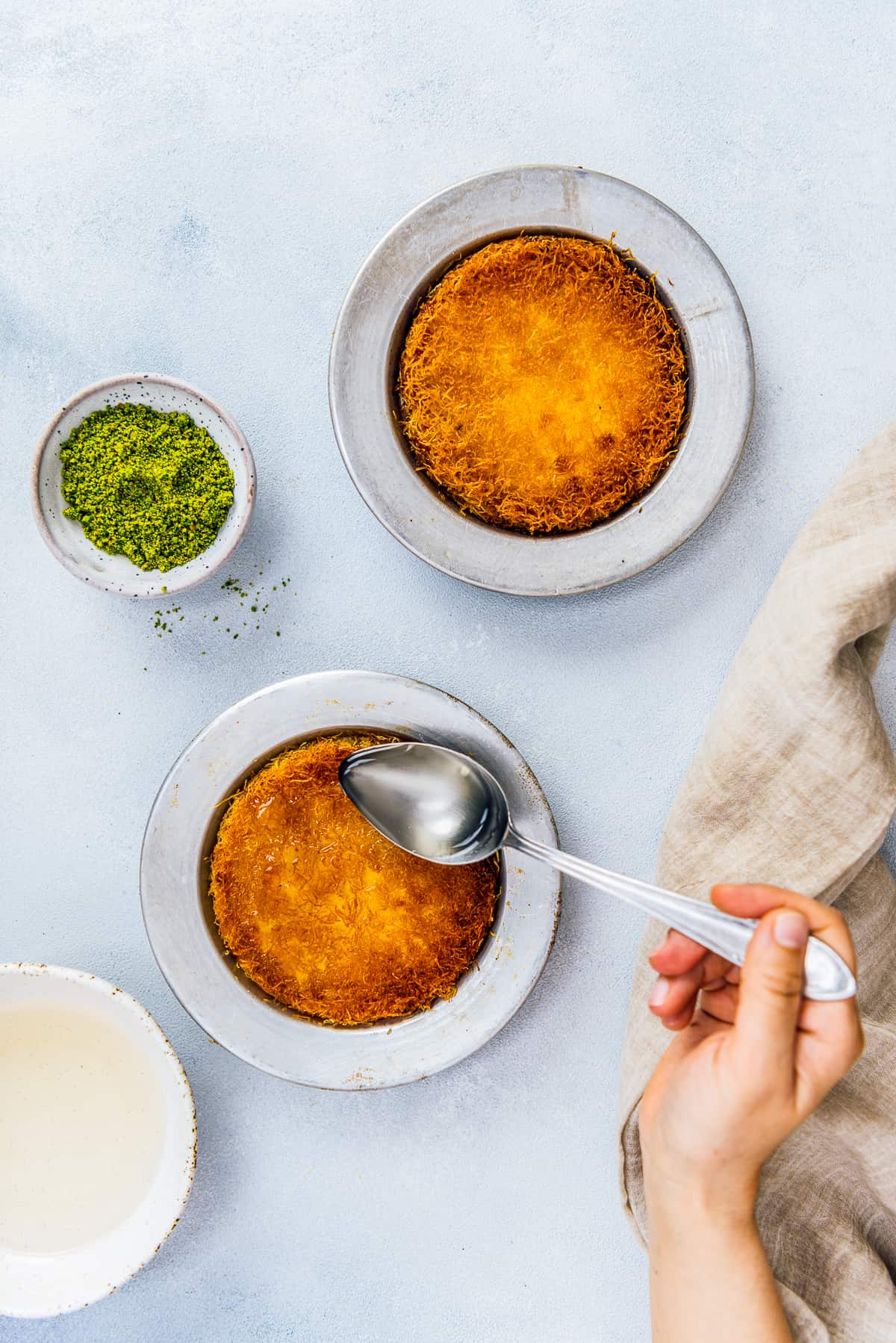 Woman drizzling syrup over Turkish kunefe in metal pans.