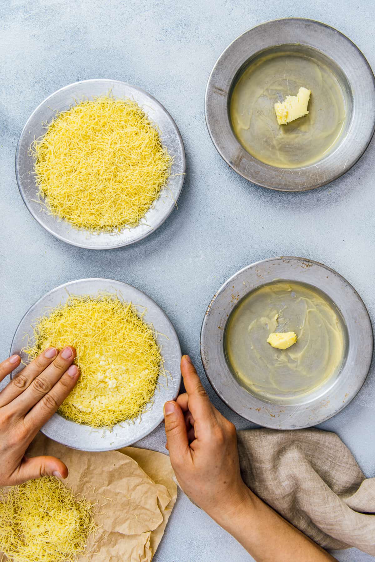 Hands placing kadayif dough and cheese in traditional metal pans.