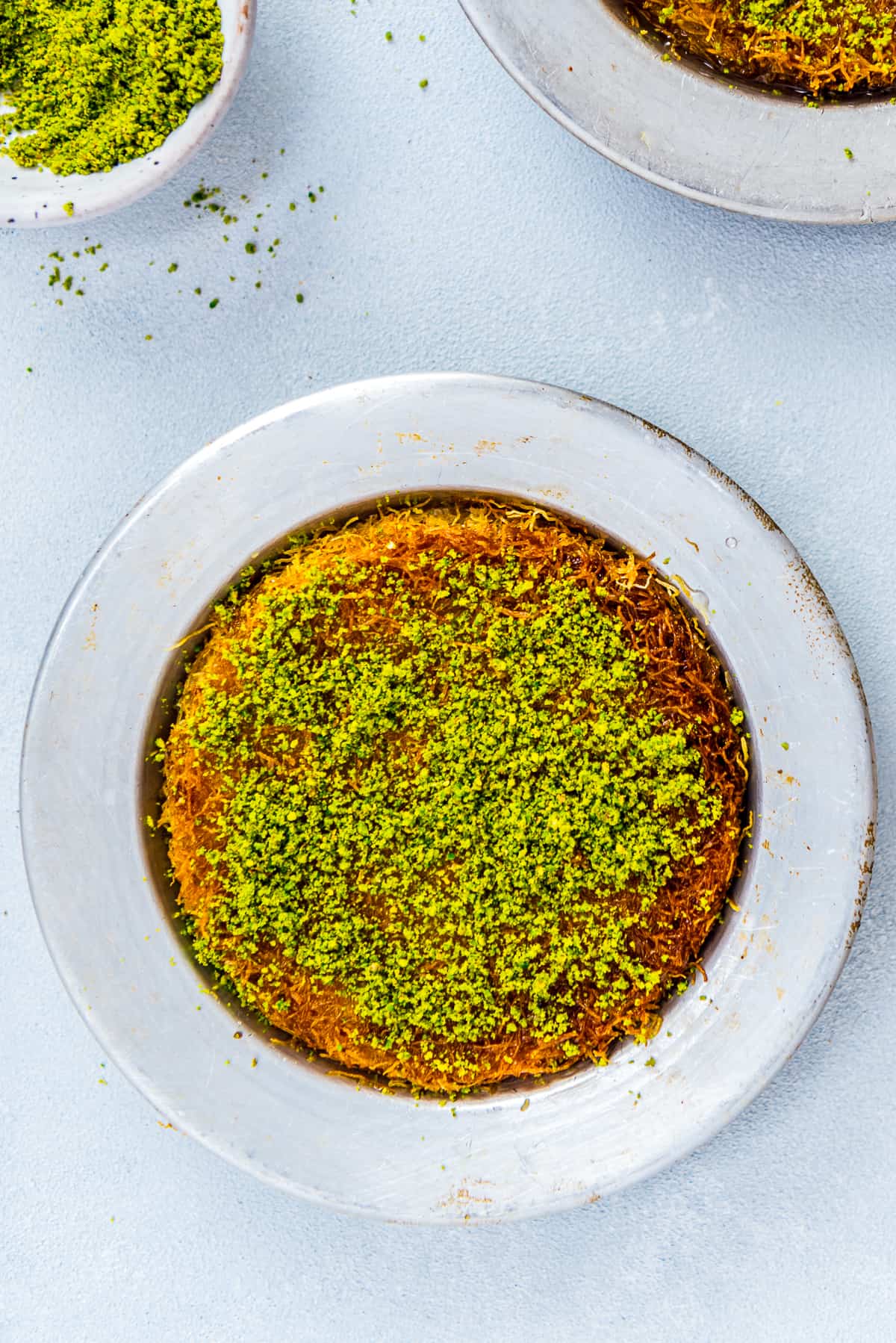 Kunefe dessert garnished with ground pistachio in a traditional metal pan.