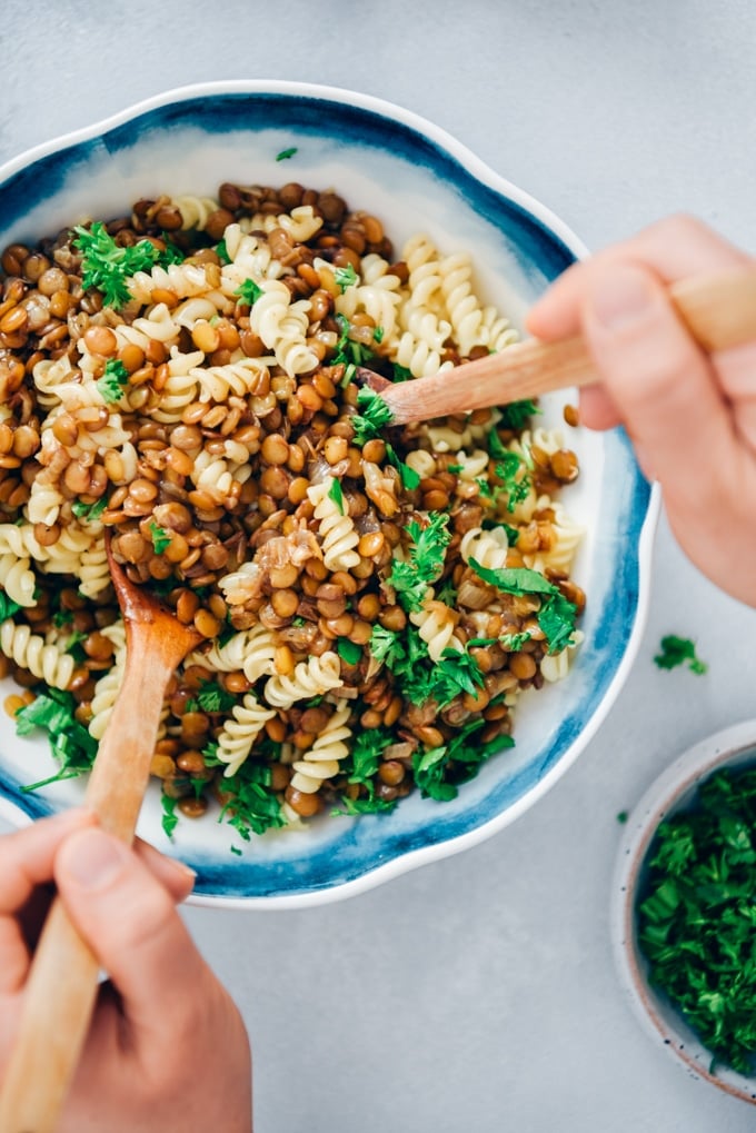 Woman stirring lentil bolognese sauce and pasta using wooden spoons.