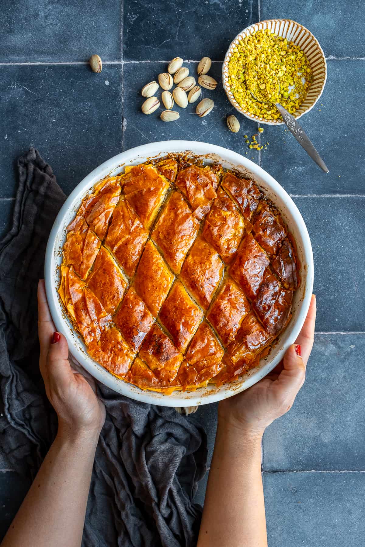 Hands holding baklava dessert in a round baking pan, pistachios and ground pistachio on the side.