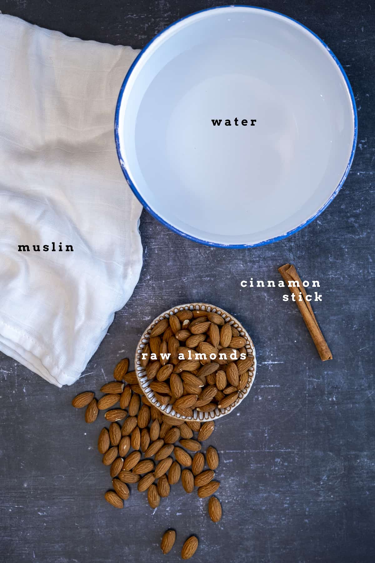 Water in a large white bowl, whole raw almonds in a bowl and on the ground, a cinnamon stick and a white muslin on a dark background.