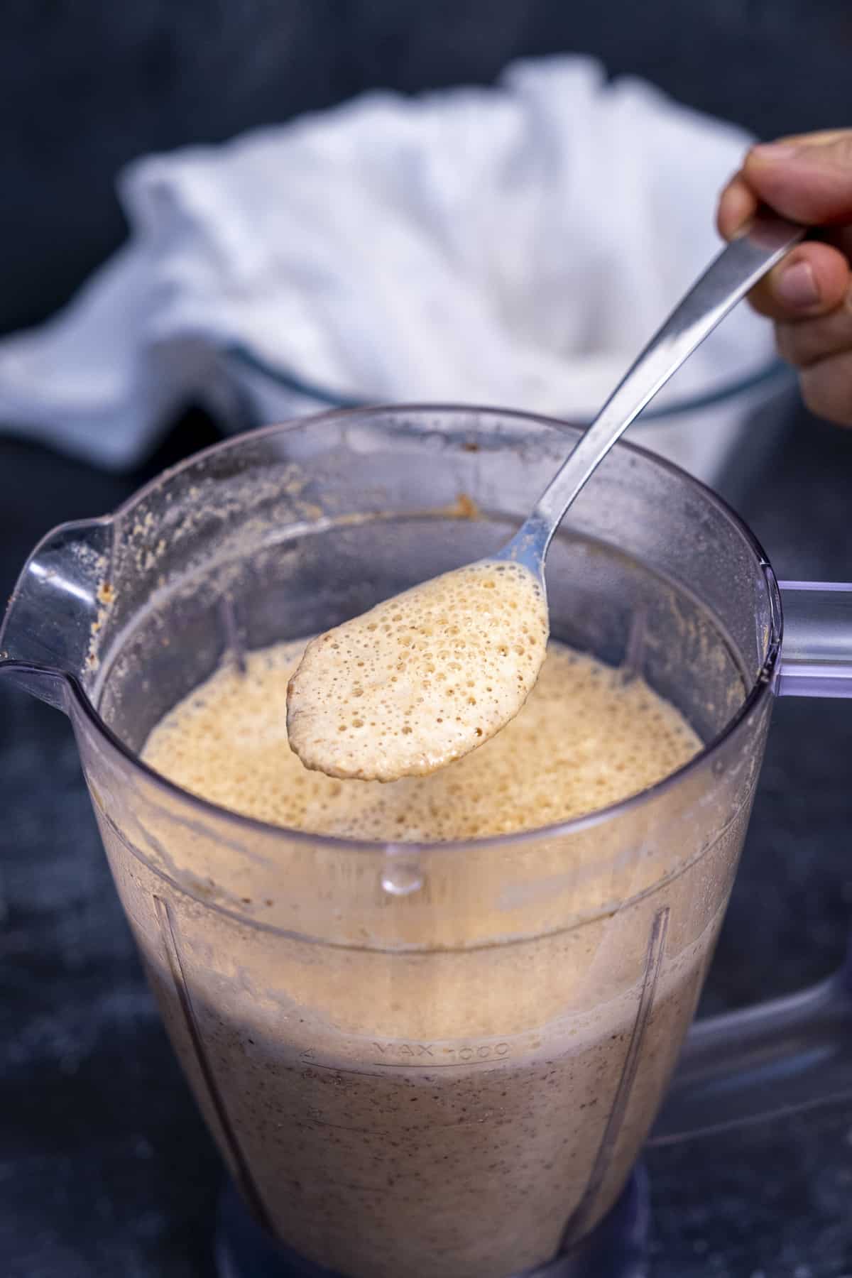 A hand holding a spoon of blended almond and water mixture over the blender container.