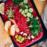 Roasted beet hummus garnished with herbs, cucumber, olives, almonds, tahini and lemon served on a black plate.