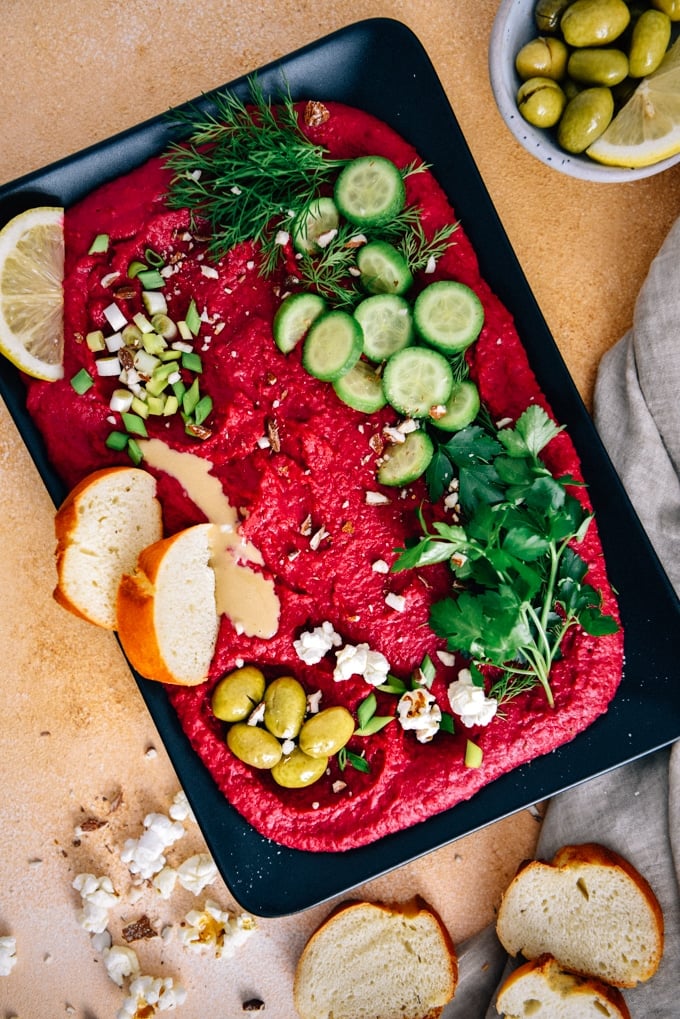 Roasted beet hummus garnished with herbs, cucumber, olives, almonds, tahini and lemon served on a black plate.