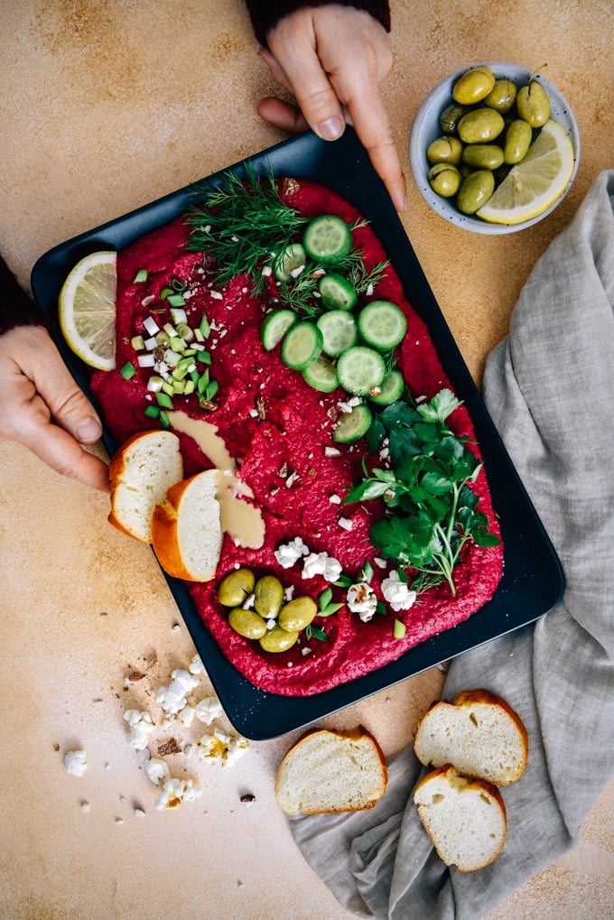 Serving beetroot hummus garnished with herbs, cucumber, olives, almonds, tahini and lemon served on a black plate.