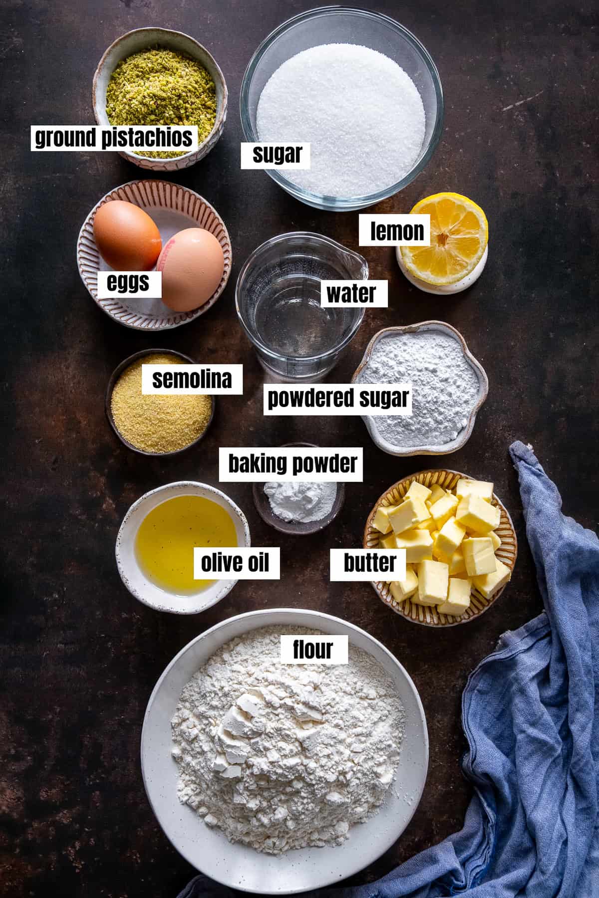 Flat lay of Şekerpare ingredients including flour, butter, olive oil, semolina, powdered sugar, eggs, lemon, water, sugar, baking powder, and ground pistachios, all labeled.