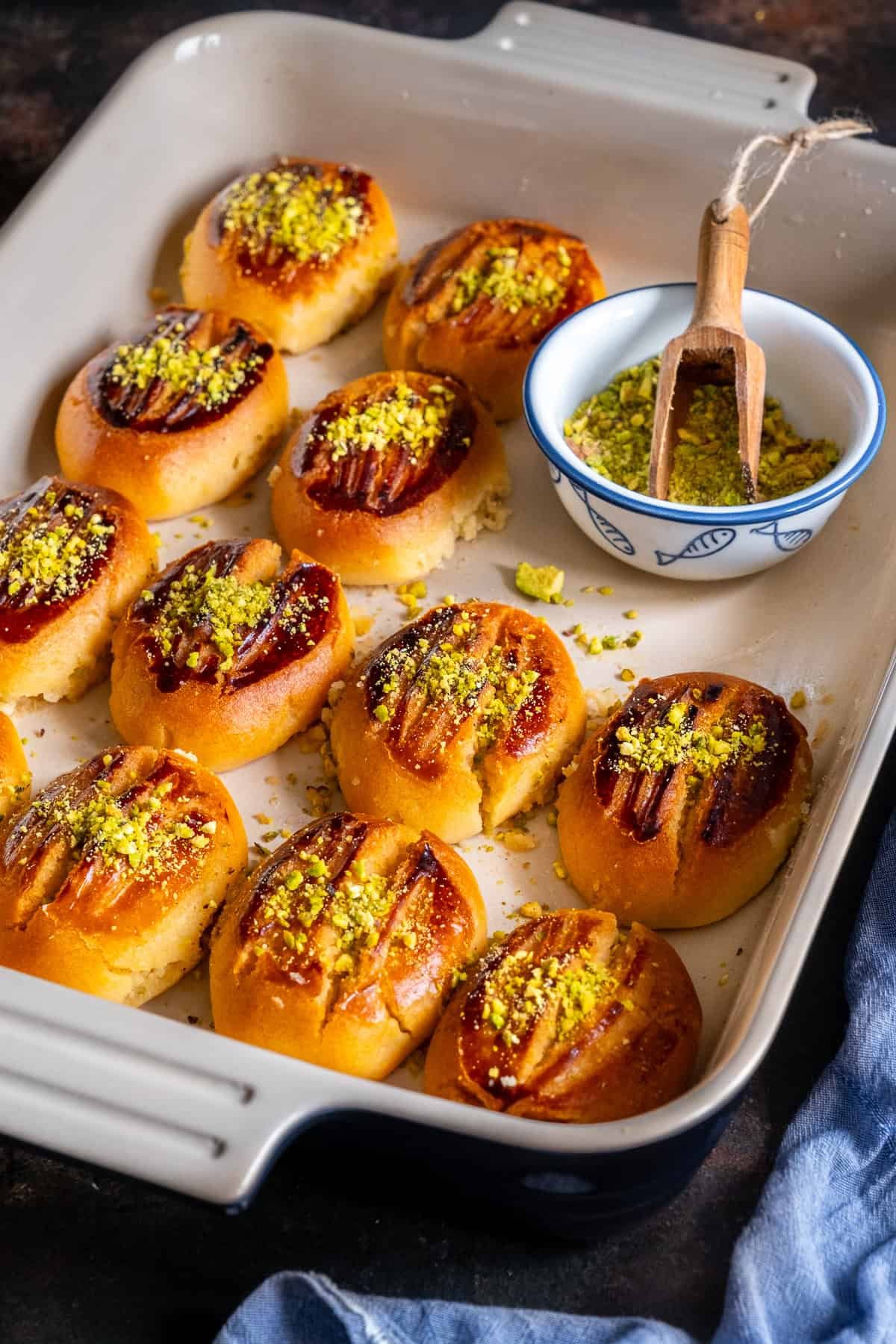 Baked Turkish Şekerpare cookies arranged in a baking dish, topped with ground pistachios, with a bowl of extra pistachios and a small wooden scoop on the side.