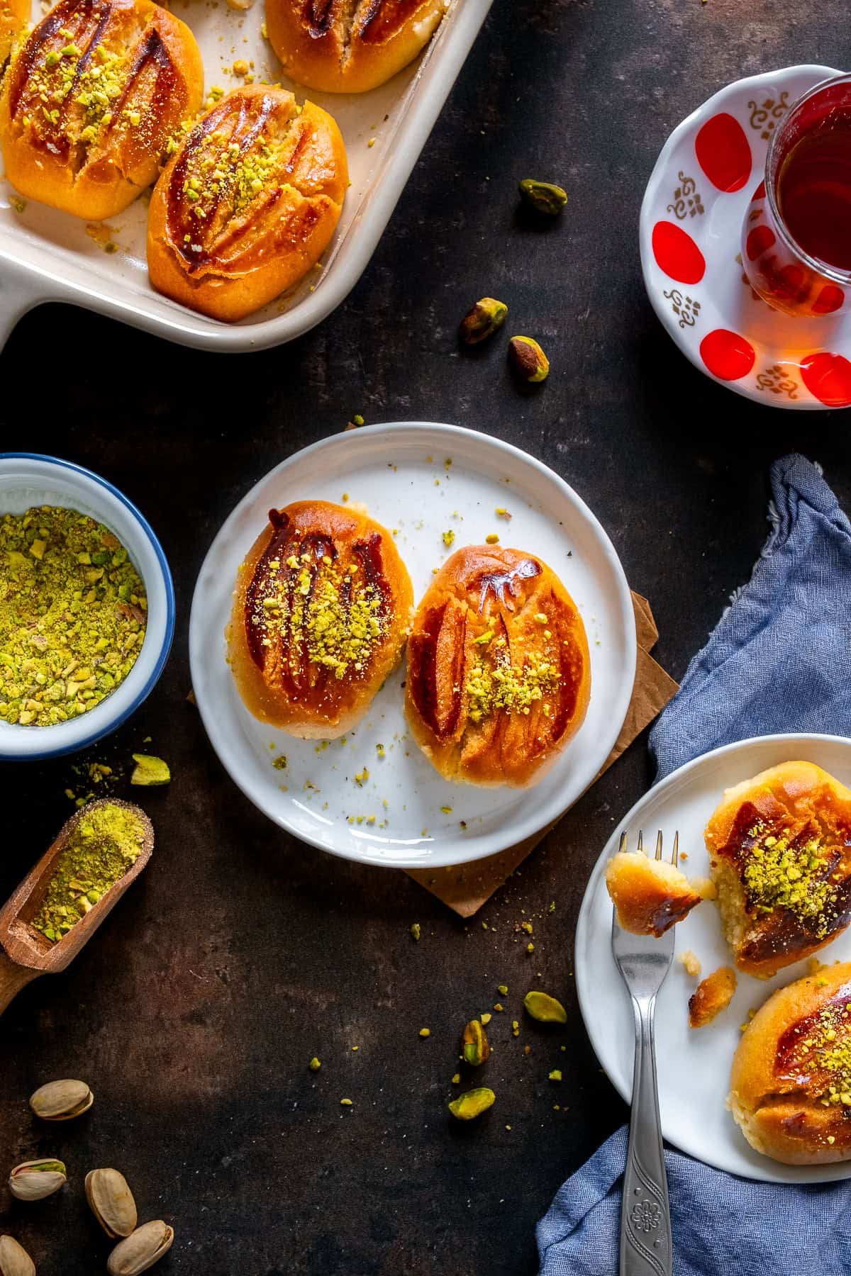 Turkish Şekerpare cookies served on small plates, topped with ground pistachios, with a glass of Turkish tea and crushed pistachios on the side.
