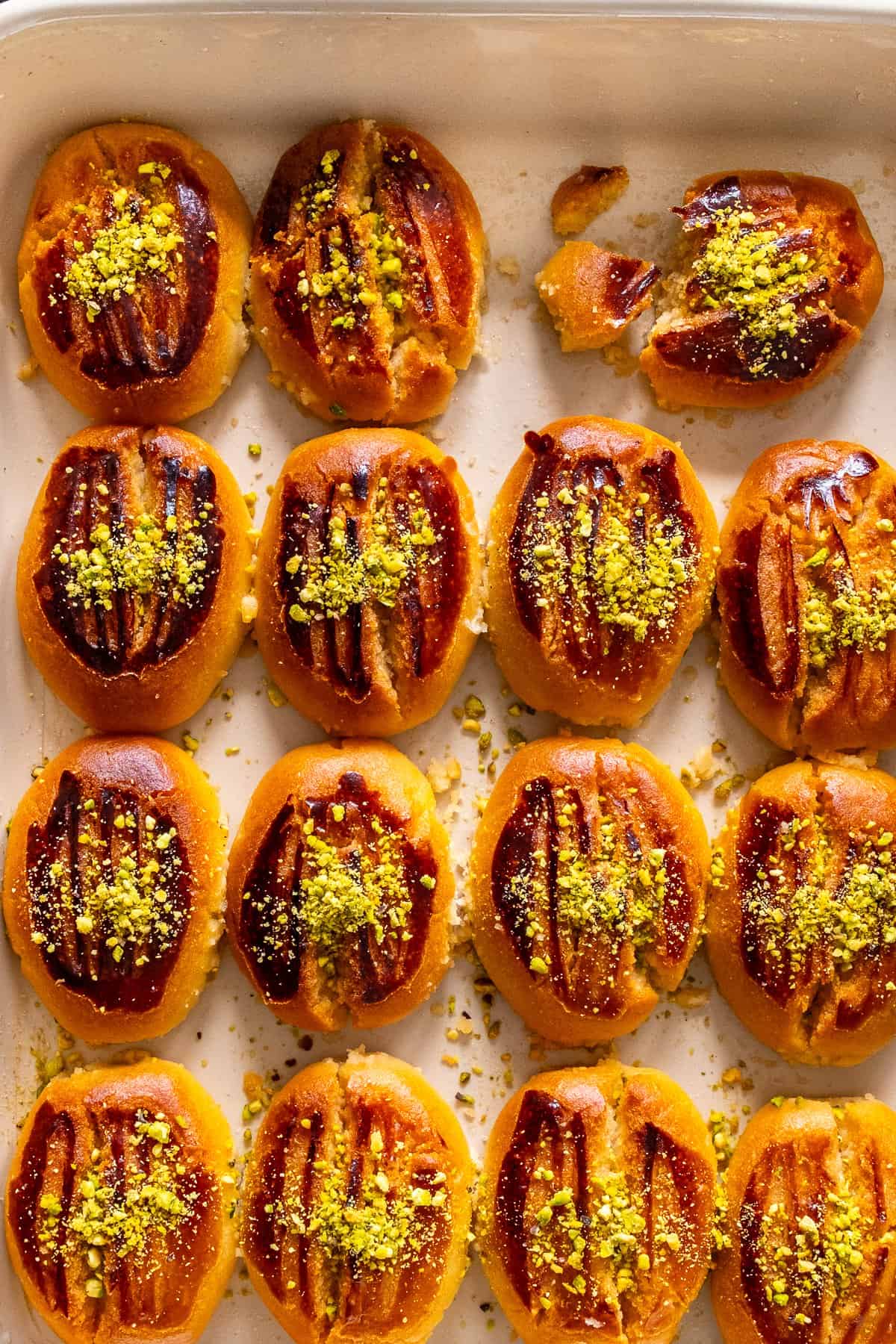 Close-up of baked Şekerpare cookies in a white baking dish, topped with ground pistachios, with one cookie broken to show its soft texture.