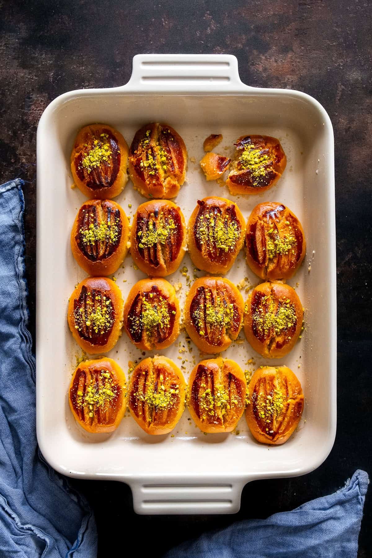 Top view of freshly baked Şekerpare cookies in a white baking dish, topped with ground pistachios, with a blue cloth on the side.