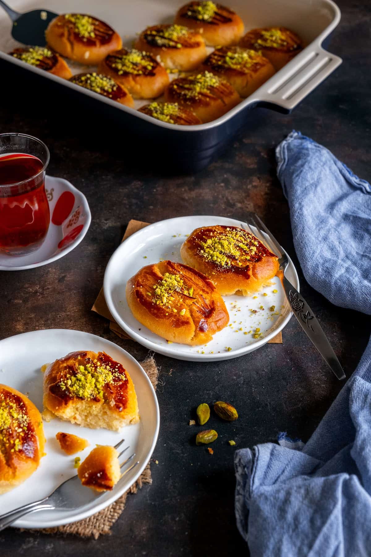 Plated Turkish Şekerpare cookies topped with ground pistachios, served with a glass of tea, and more cookies in a baking dish in the background.