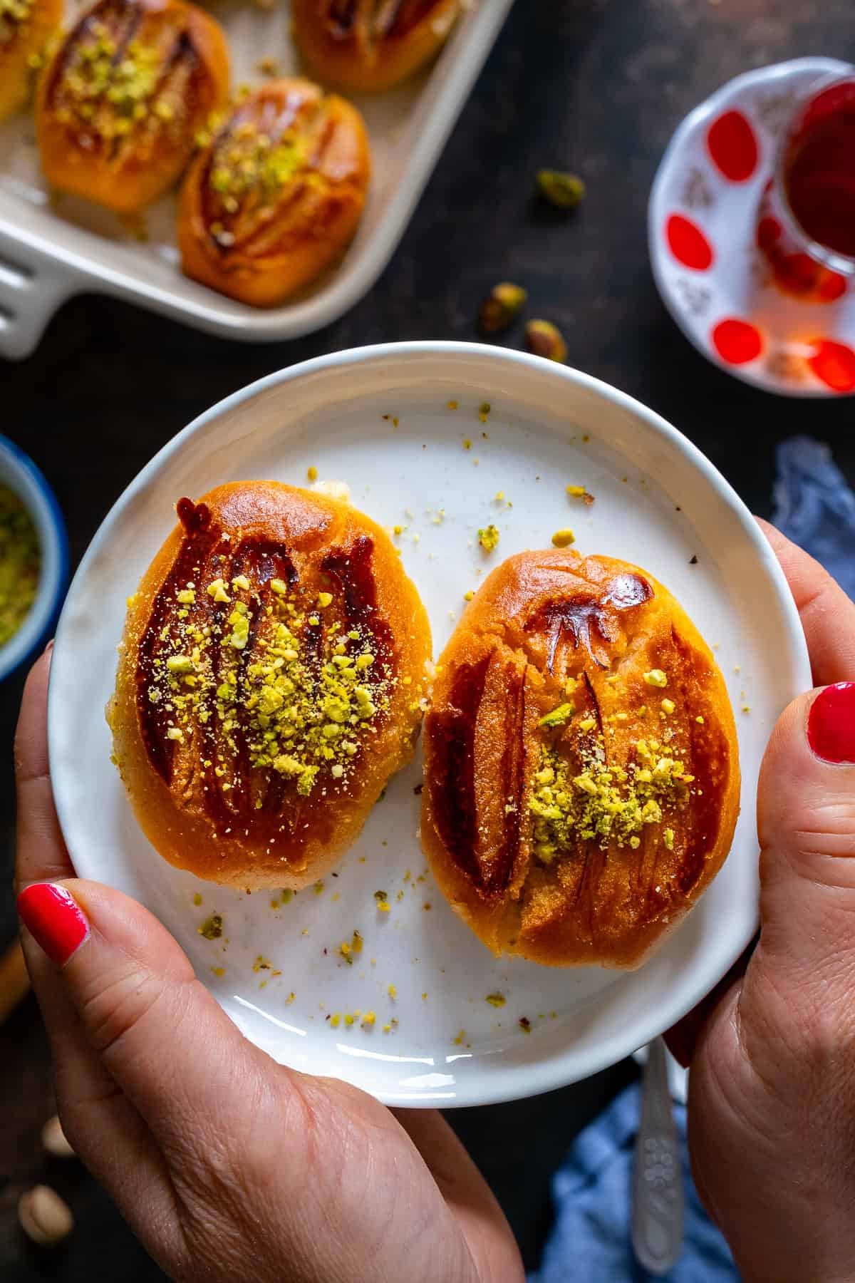 Two Turkish Şekerpare cookies topped with ground pistachios on a small white plate, held in hands with red nail polish.