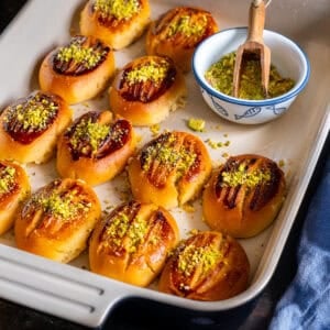 Freshly baked Turkish Şekerpare cookies in a baking dish, topped with ground pistachios, with a small bowl of extra pistachios on the side.