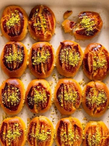 Golden brown Şekerpare cookies arranged in a white baking dish, topped with ground pistachios, with one broken to show the soft inside.