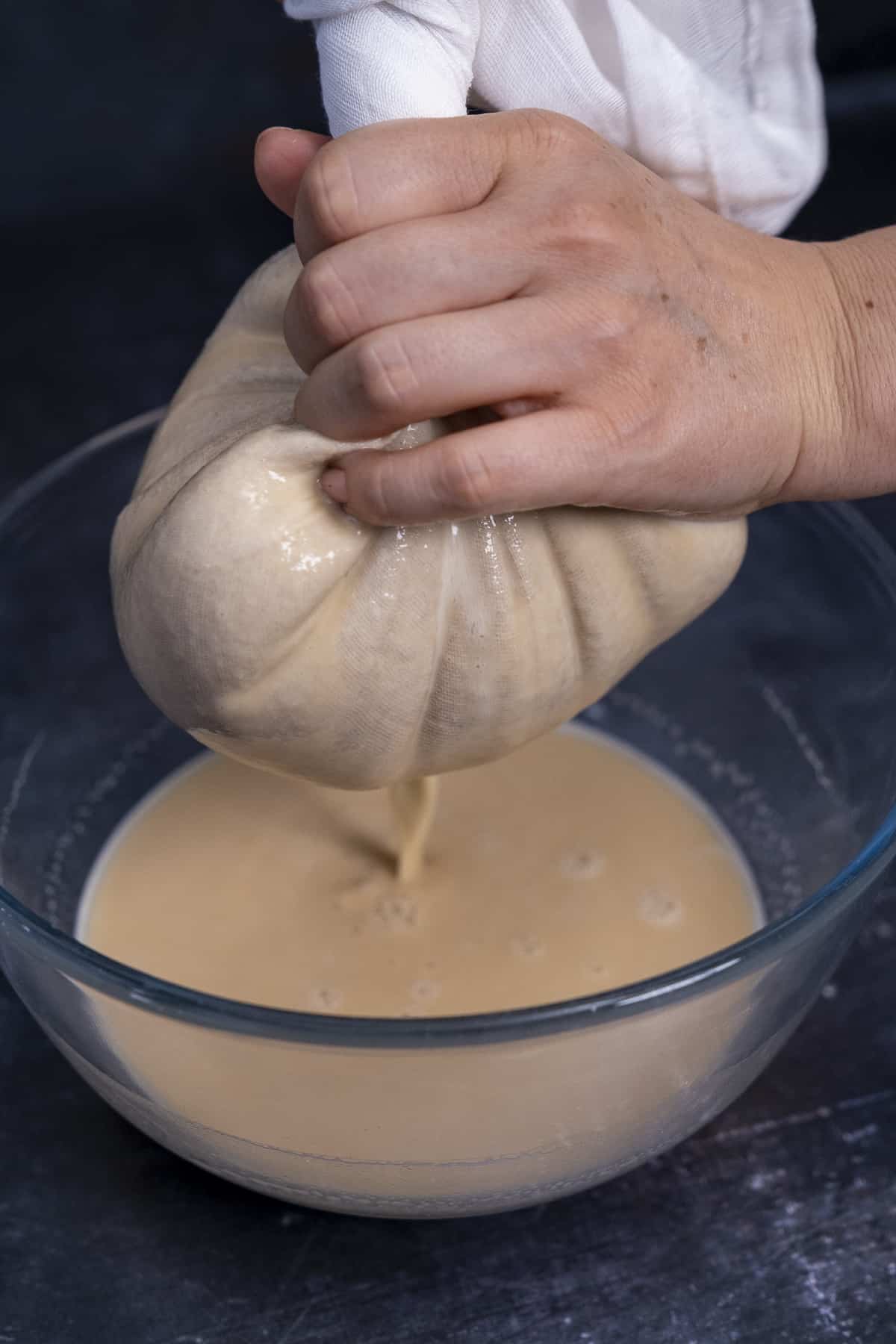 Hands straining almond milk in a white muslin into a large bowl.
