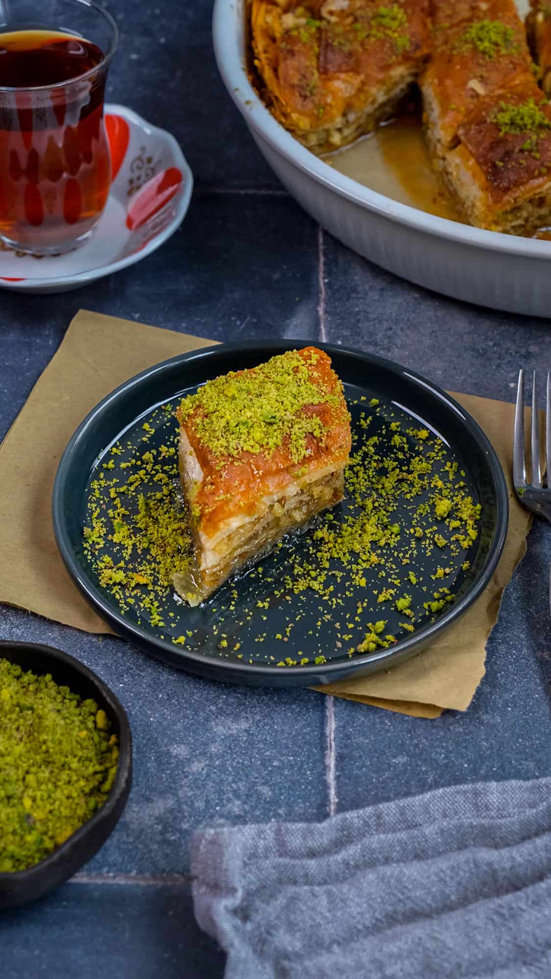 A slice of baklava garnished with ground pistachio served on a dark grey plate, tea in a glass and more baklava in a pan on the side.