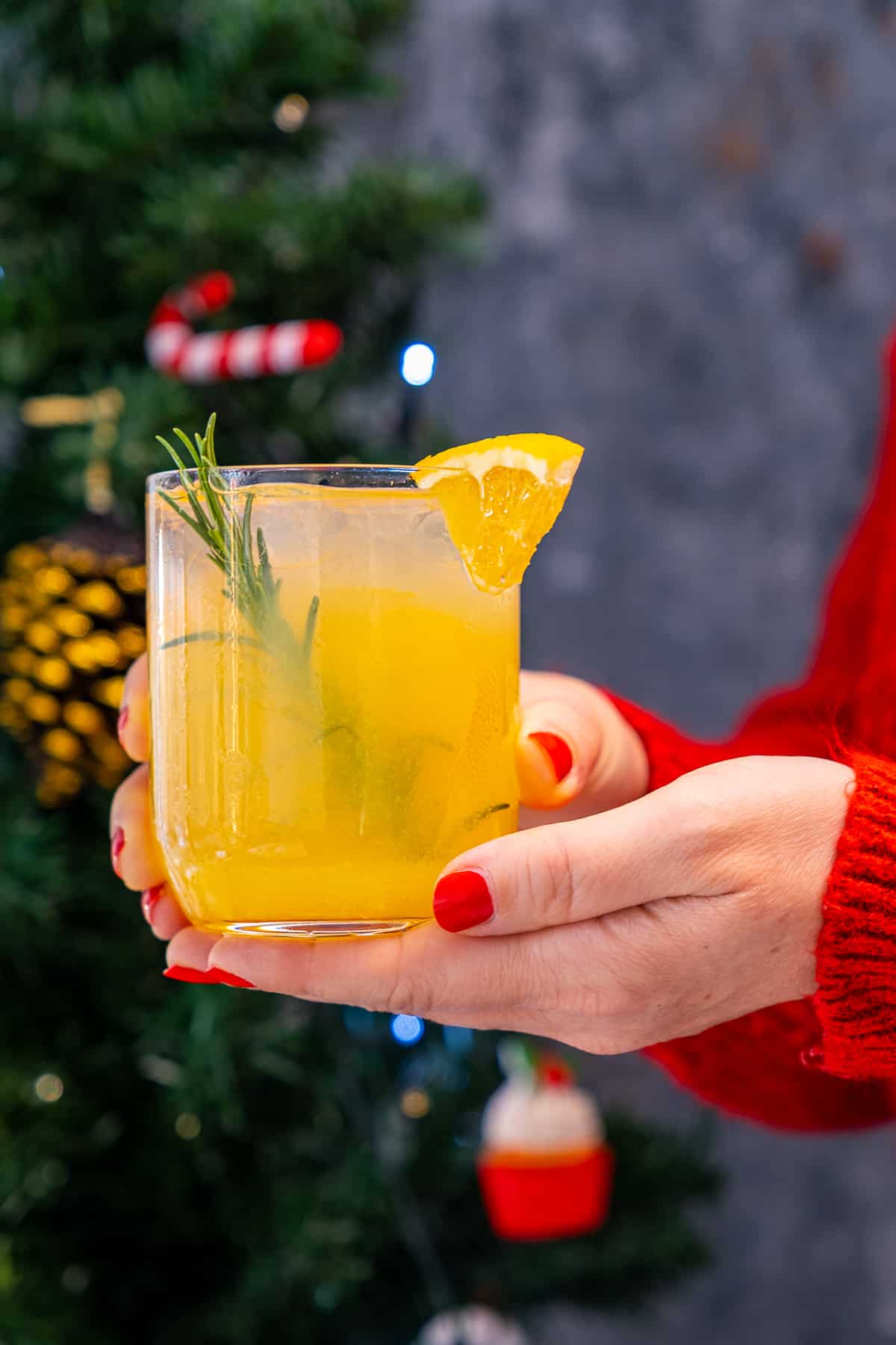 Woman with a red jumper holds a glass of vodka orange cocktail and a Christmas tree on the background.